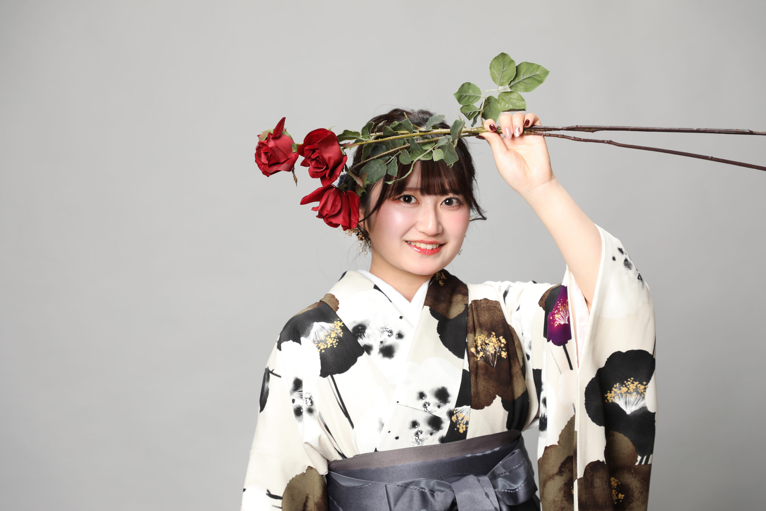 赤い薔薇を頭上にかざしていたずらっぽく笑う椿柄の着物と袴の女性