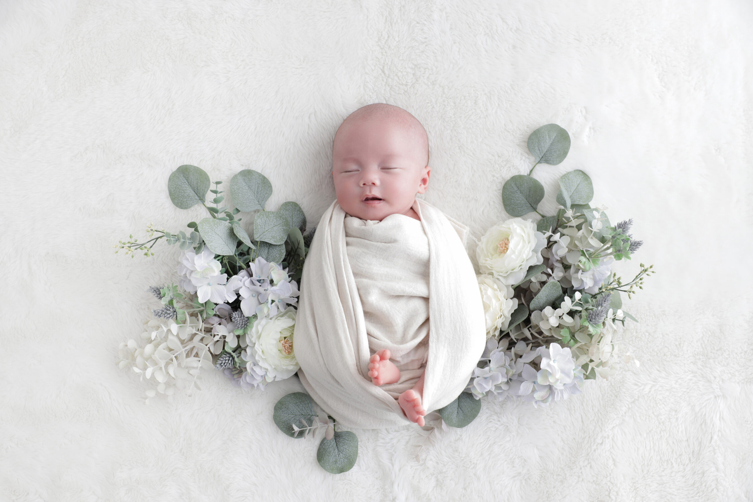 白いおくるみに包まれユーカリと花に囲まれて眠る新生児