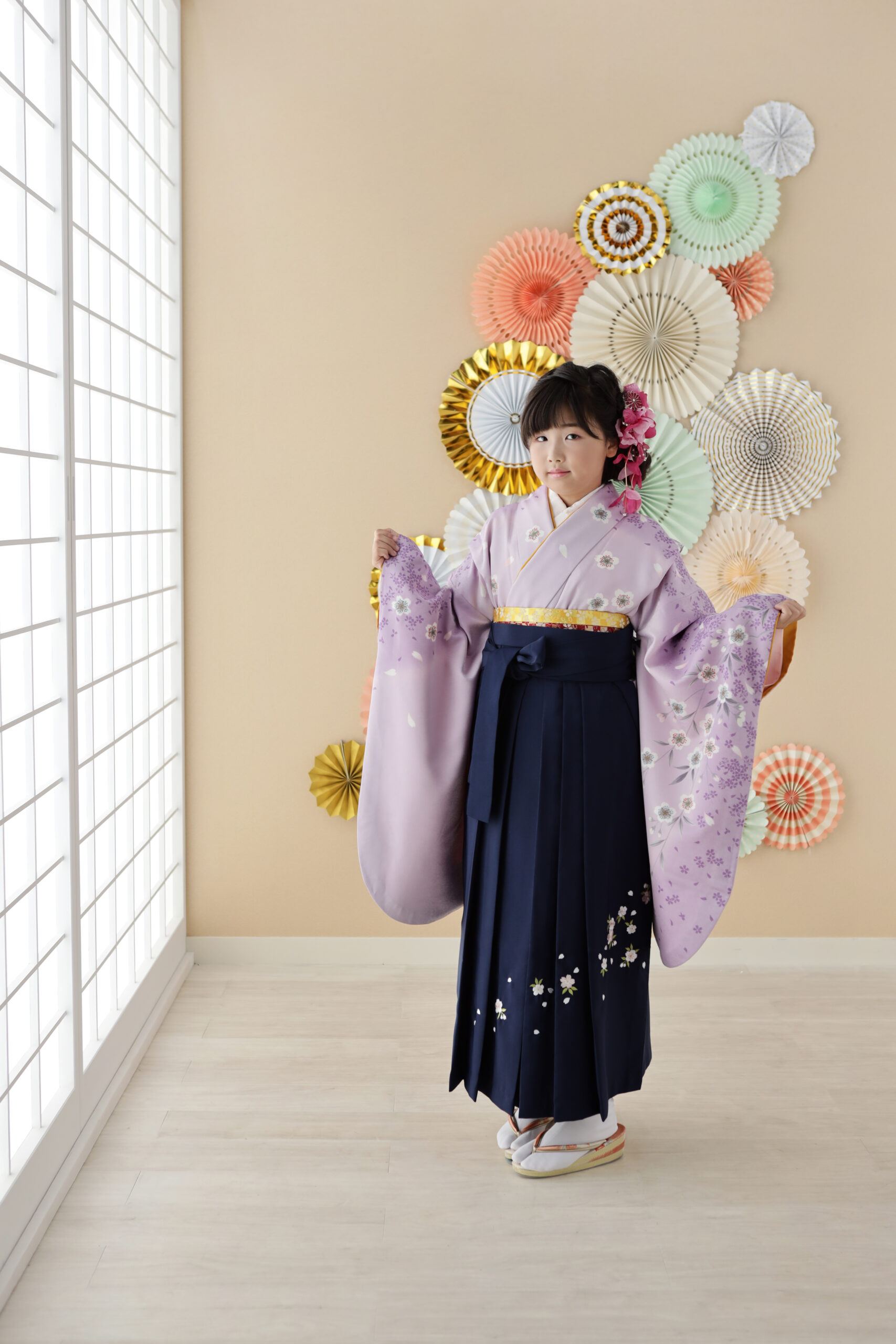 ペーパーファンを飾った和室で薄紫の着物の袖を広げる十歳の女の子