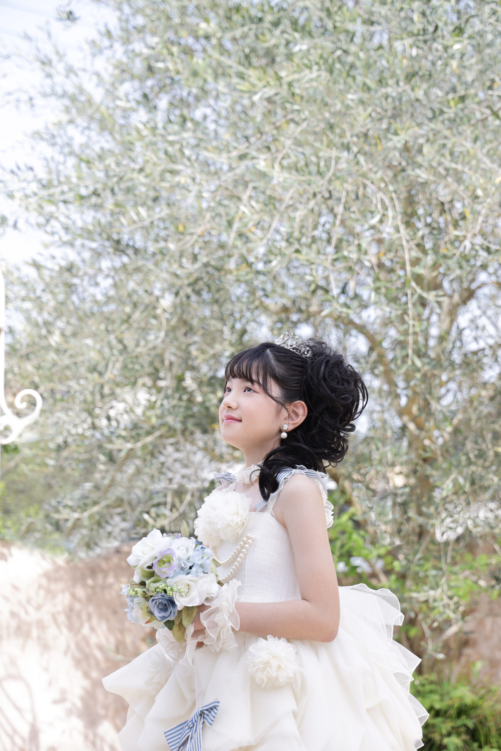 オリーブの木の下でティアラを着け白いドレスでブーケを抱える女の子
