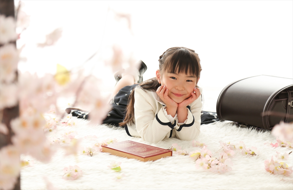 桜の枝の向こうで頬杖をつき洋書とランドセルのそばに寝ころぶ女の子