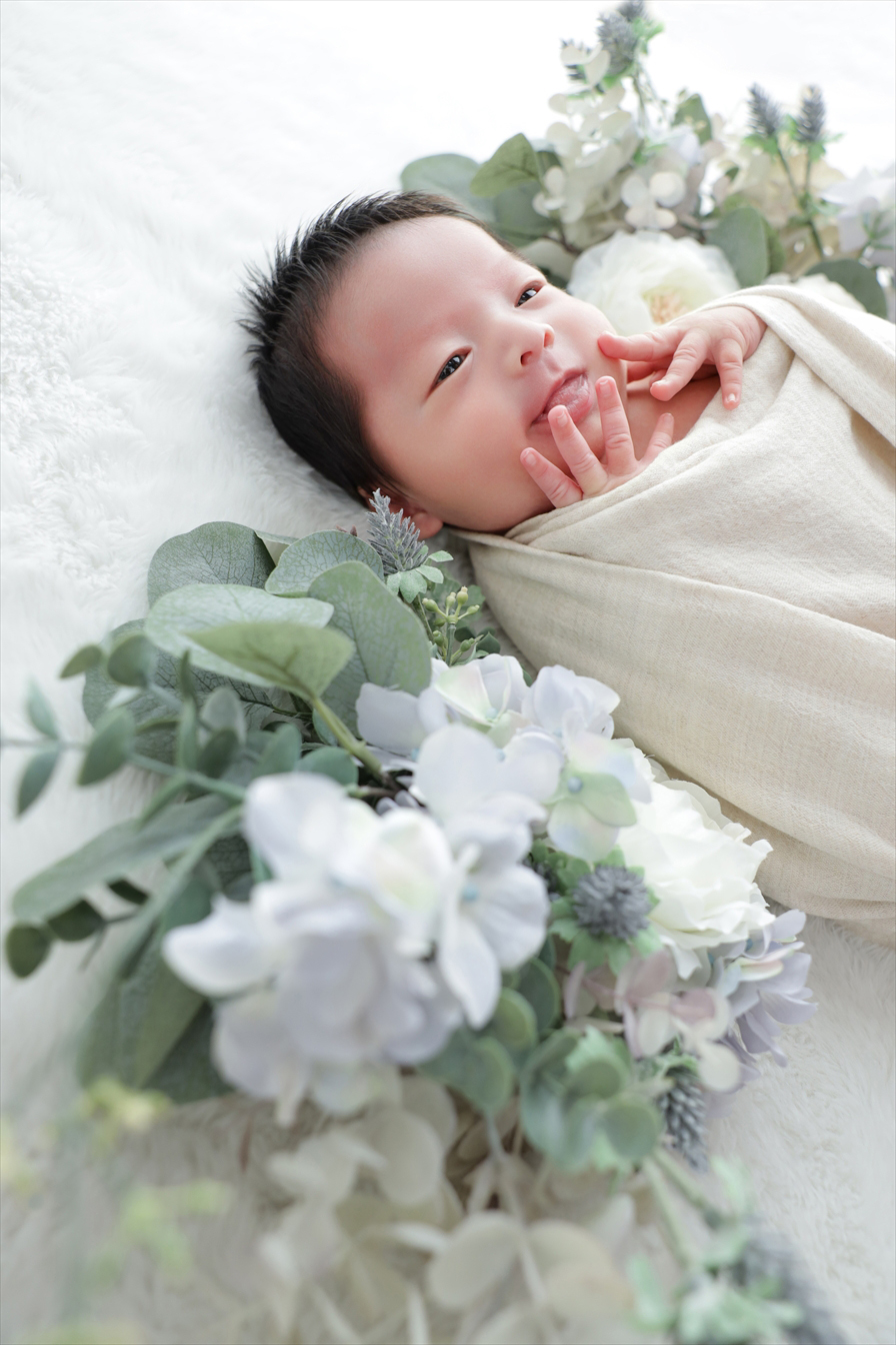 生成りのおくるみ姿で口元に手を添え目を開ける新生児