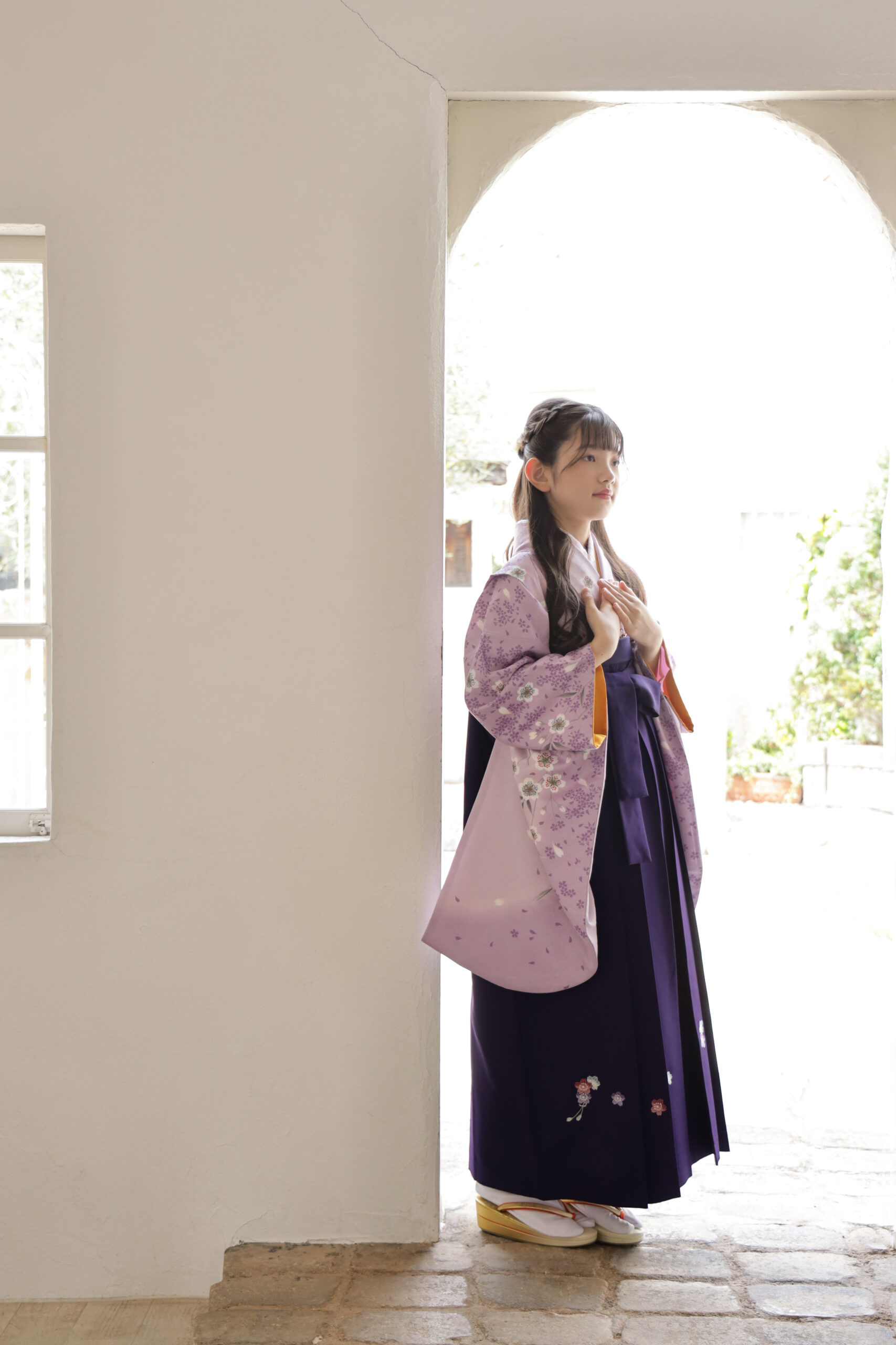 アーチ形の窓辺で胸に手を添える薄紫の着物と紫袴の女の子