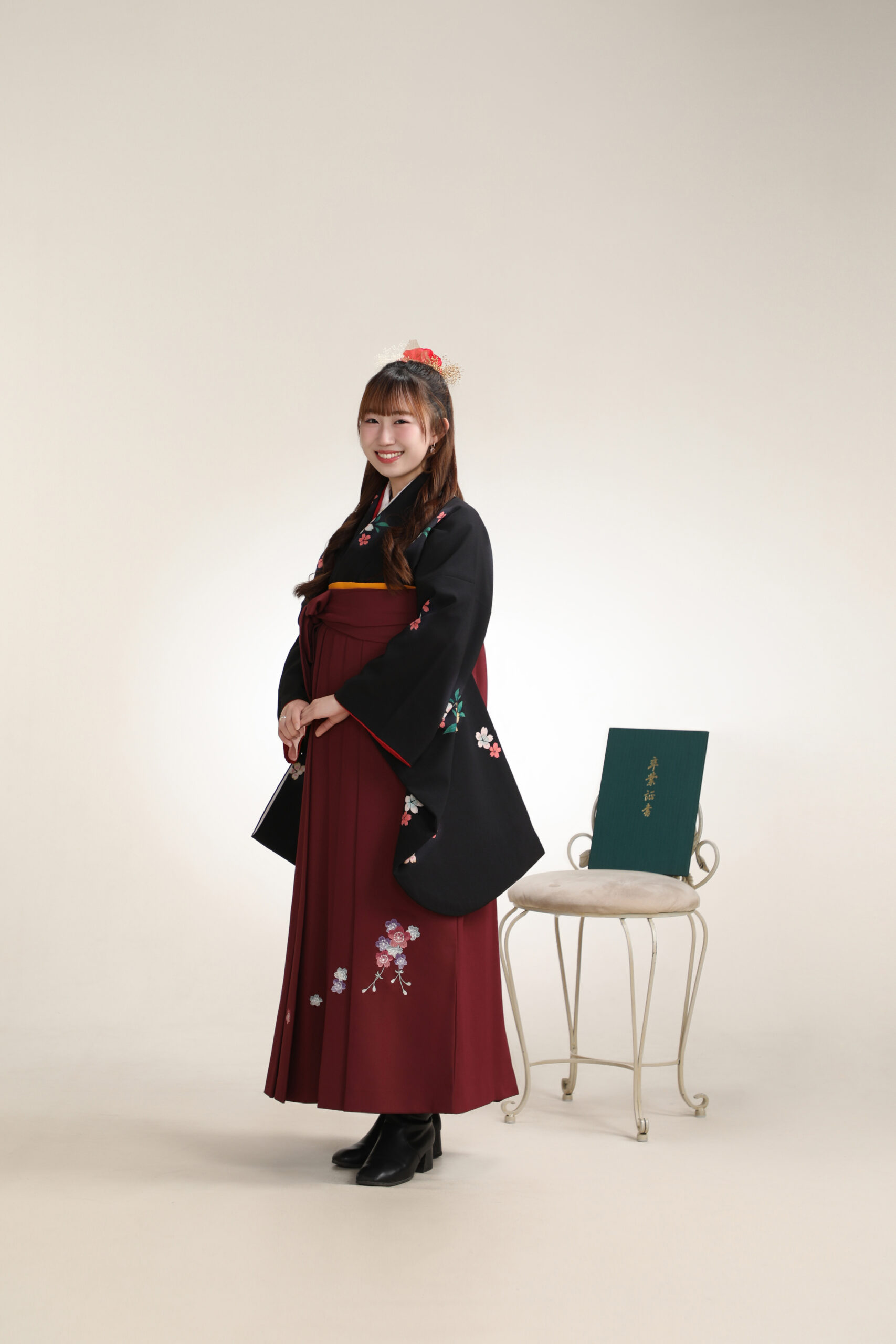 卒業証書を椅子に添え桜柄の黒い着物とえんじ袴で口元をほころばせる