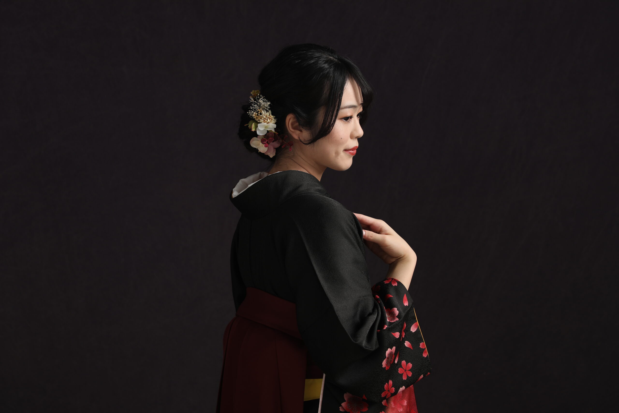 暗い背景で袖の桜に指をかけ横顔を見せる黒着物とえんじ袴の女性