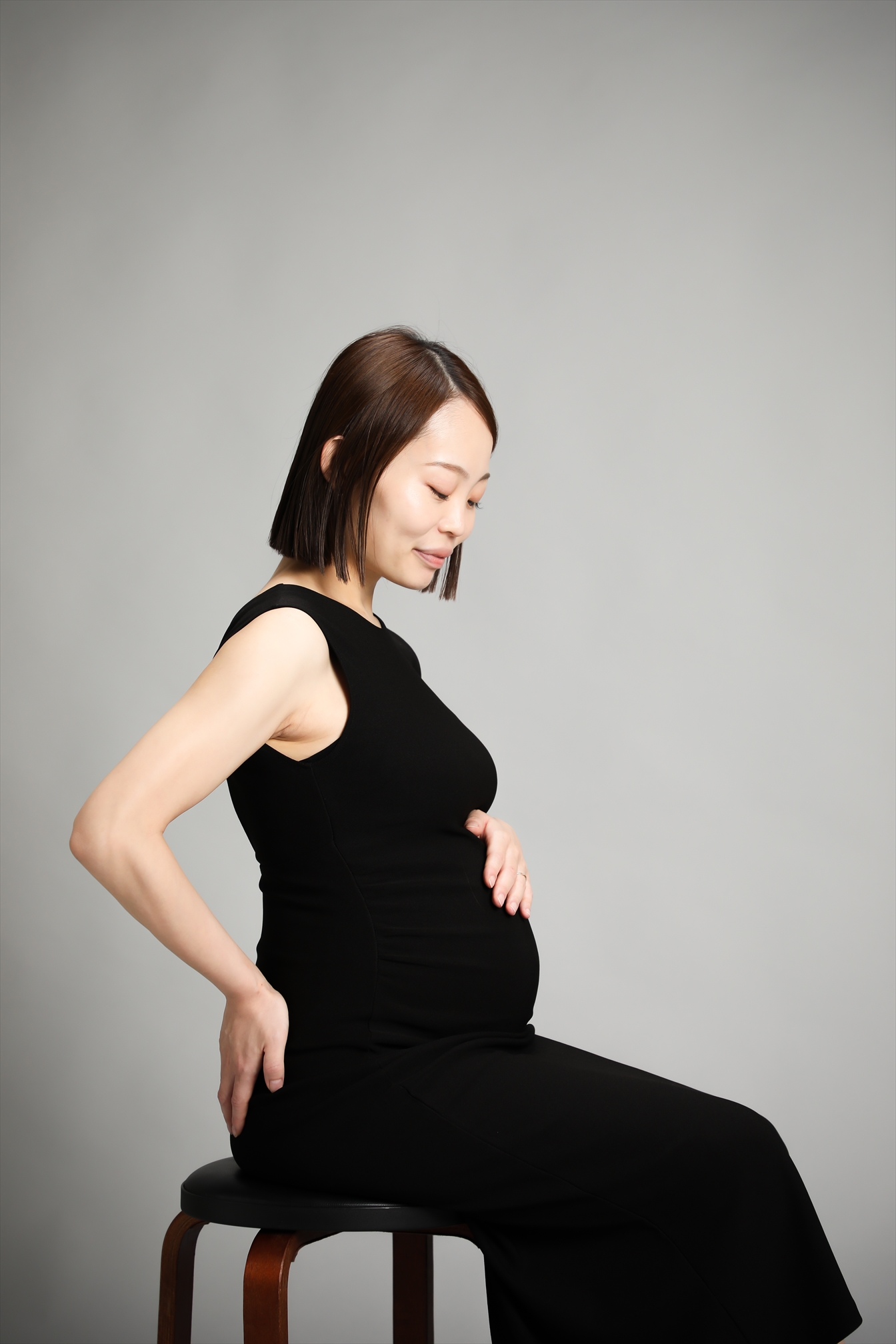 黒いドレスでスツールに腰かけおなかに手を添える横向きの姿