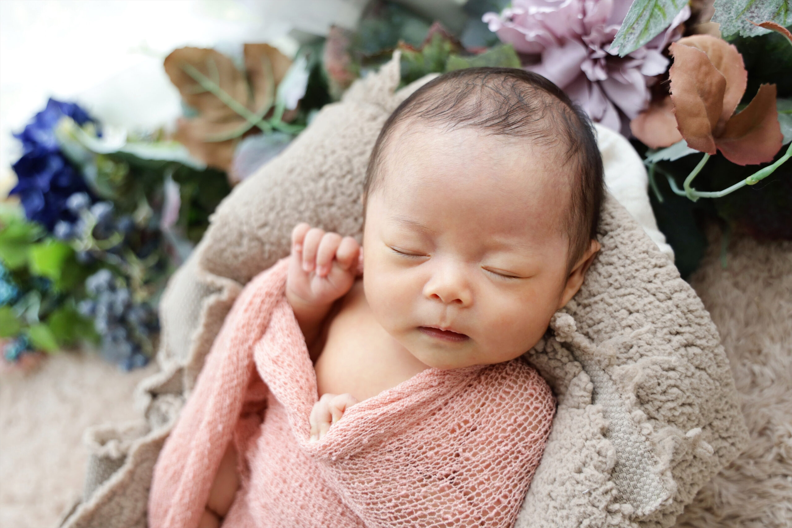 ピンクのおくるみで花々に囲まれ穏やかに眠る新生児
