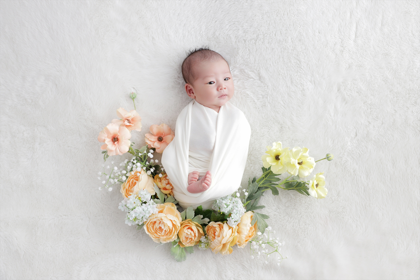 オレンジと黄色の花を三日月形に並べた中で目を開ける新生児
