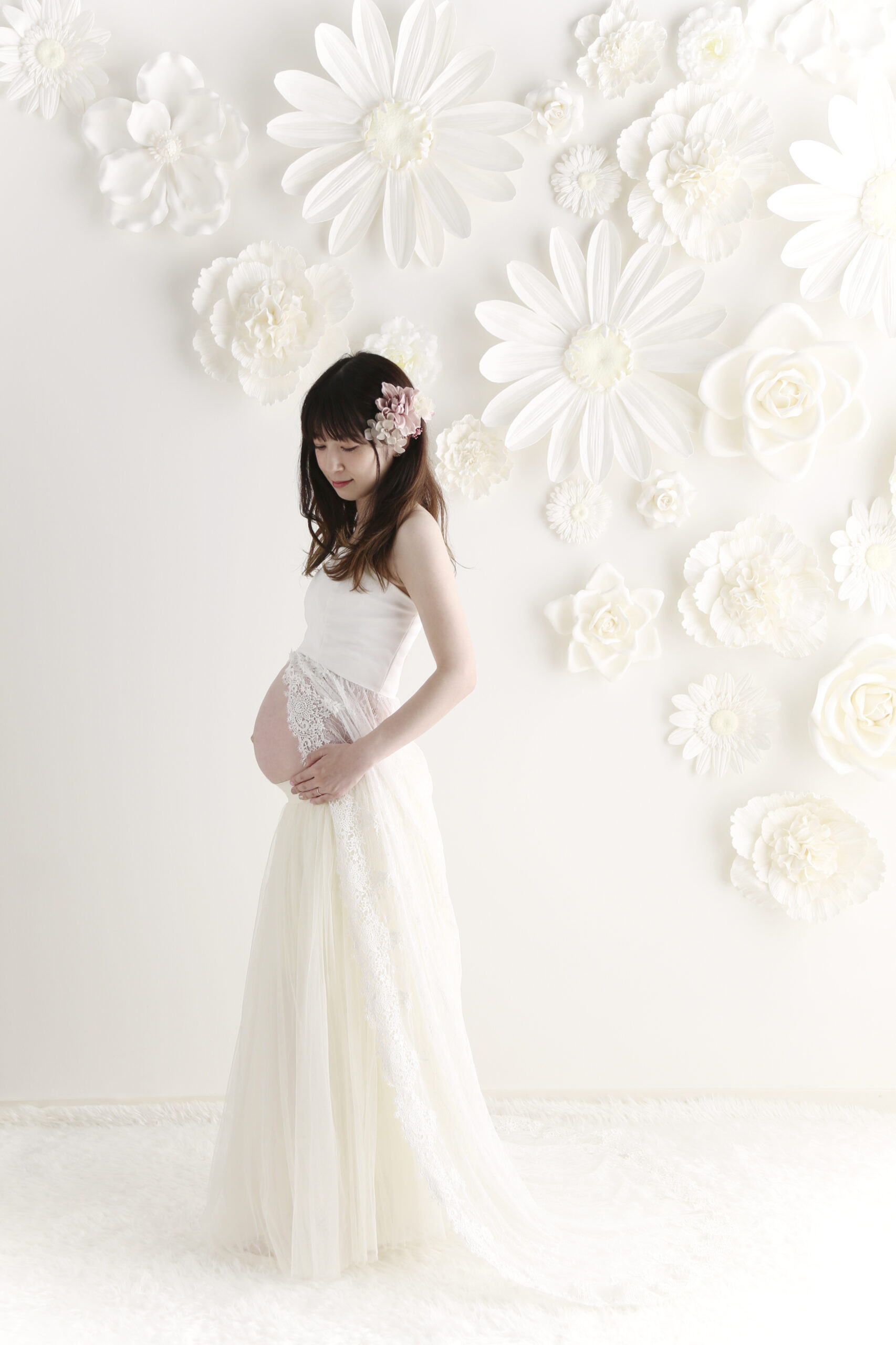 白い花を敷きつめた壁の前でおなかに手を添えるドレス姿の妊婦