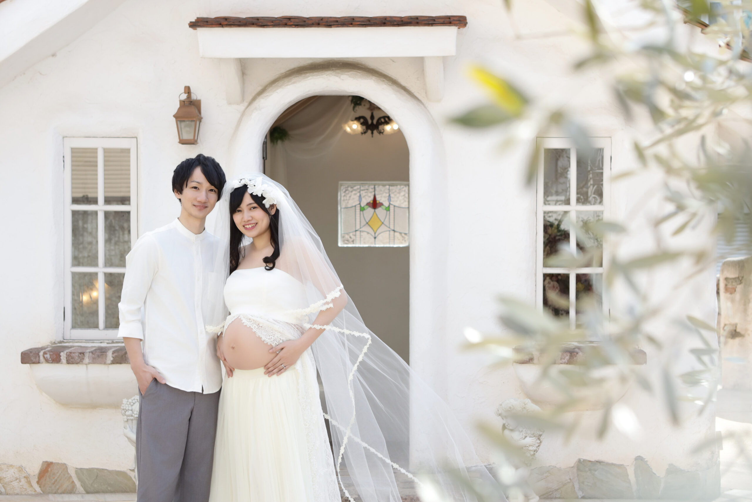 白い洋館の玄関前でベールをまといおなかに手を重ねるご夫婦
